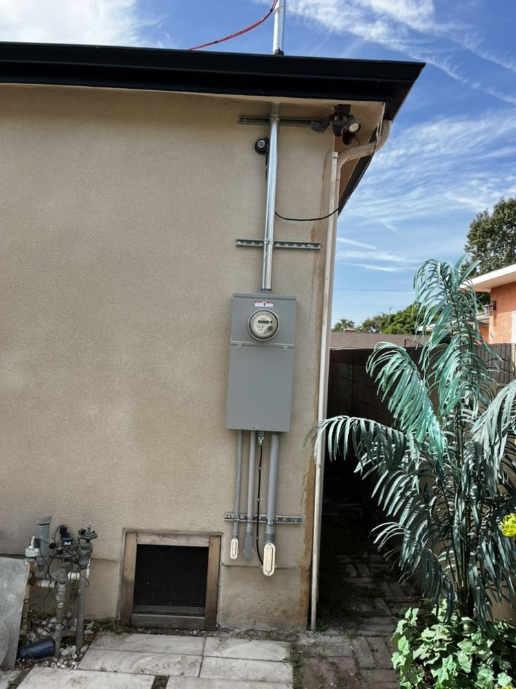 An exterior electrical meter and service panel mounted on a beige stucco wall next to a small palm tree and a crawl space.