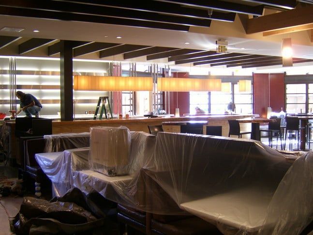 A restaurant interior under renovation, featuring covered furniture, a worker on a ladder, and warm pendant lighting.
