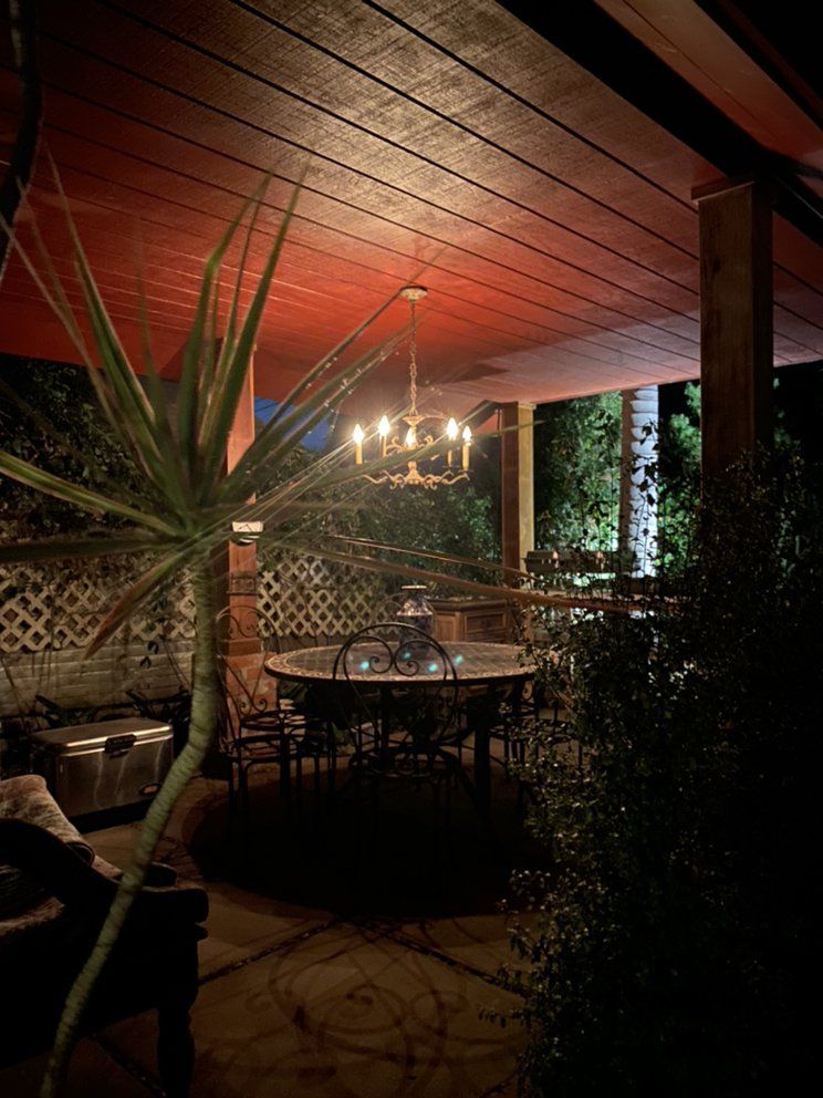 A warmly lit covered patio at night features a chandelier, dining table, and large potted plant under a wooden ceiling.
