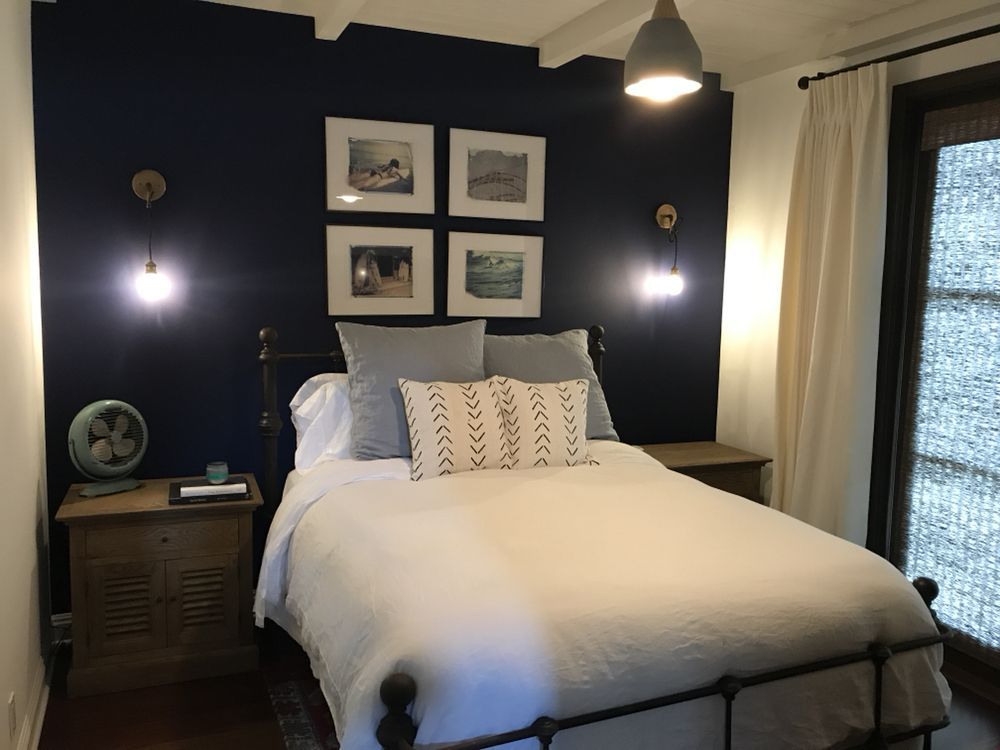 A bedroom featuring a bed with a white quilt, gray pillows, and a dark navy wall with four framed pictures and wall lights.