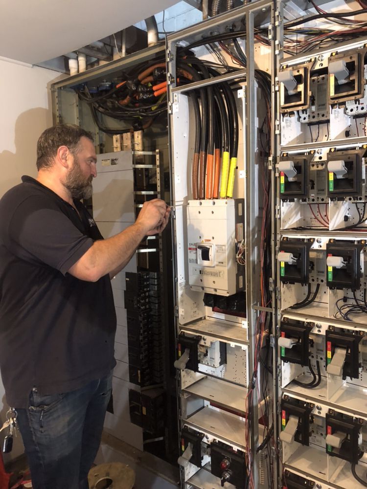 Technician working on a large open electrical distribution panel with exposed wiring and multiple circuit breakers.