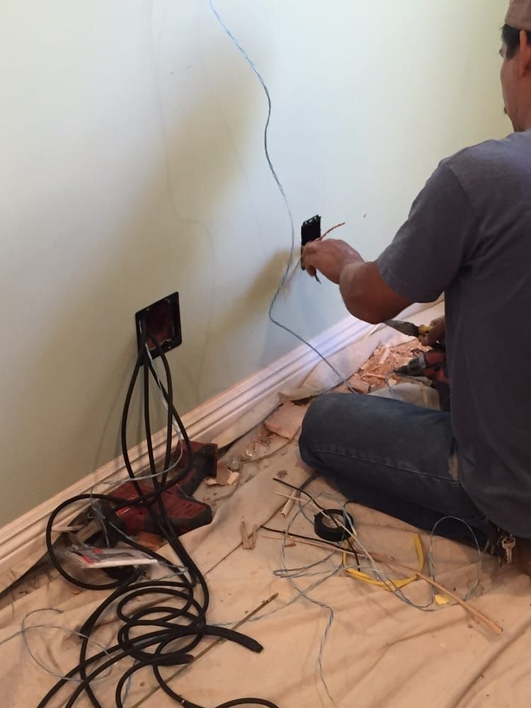 A person sits on the floor, installing electrical wiring into a wall outlet near baseboards.