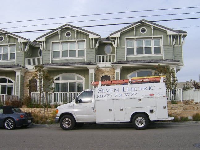 A Seven Electric service truck parked in front of residential townhouses with green siding and stone trim.