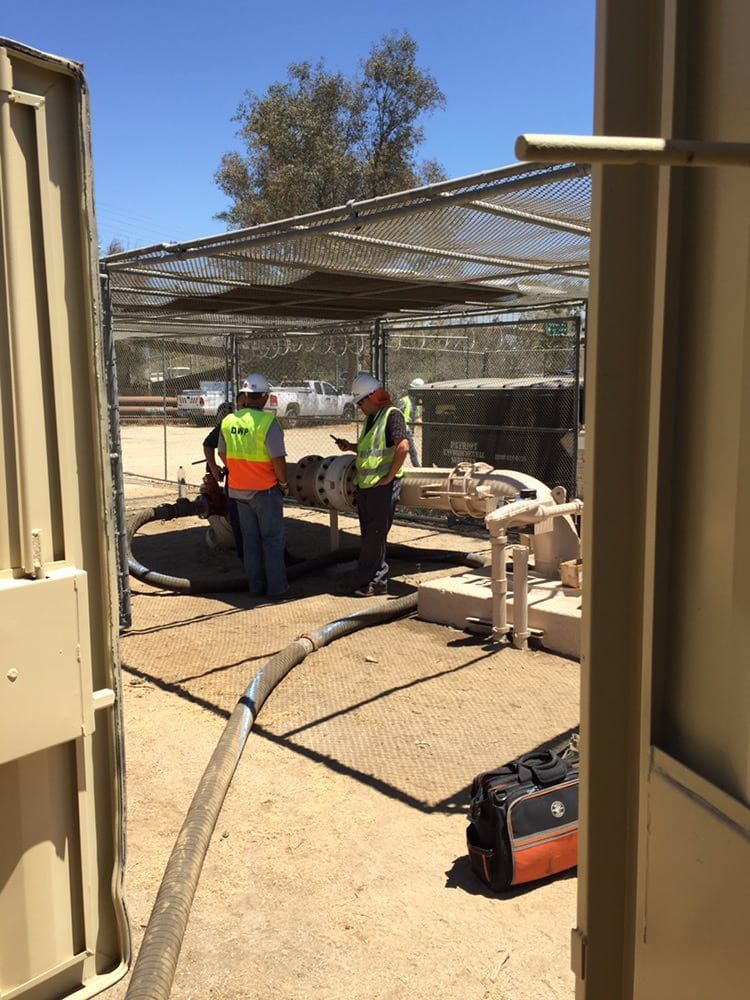 Two workers in safety vests and hard hats inspect a large pipe installation in an outdoor, fenced industrial area.