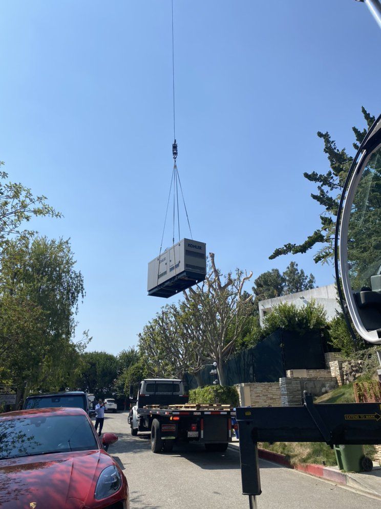 A crane hoists a large, rectangular generator over a residential street with parked cars.