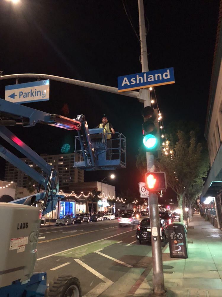 A worker in a yellow safety vest operates a blue cherry picker lift near a street sign reading