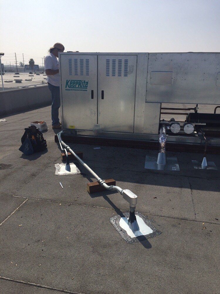A technician stands next to an HVAC unit on a rooftop, with a metal conduit running from the unit to a sealed pipe.