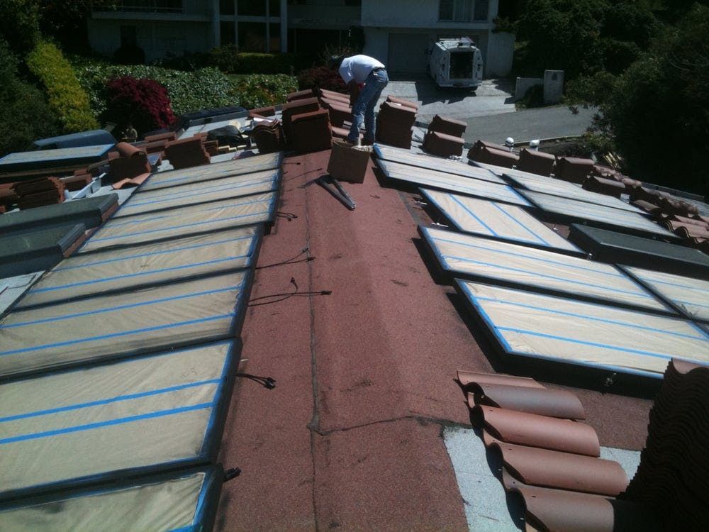 A worker in a white shirt repairs a tiled roof with several solar panels installed on the slopes.
