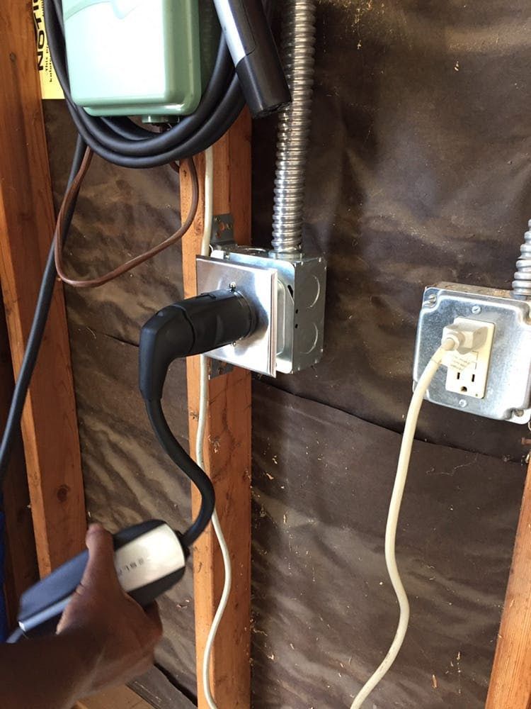 A person plugs a charging cable into an electrical outlet mounted on a wooden wall in an unfinished garage.