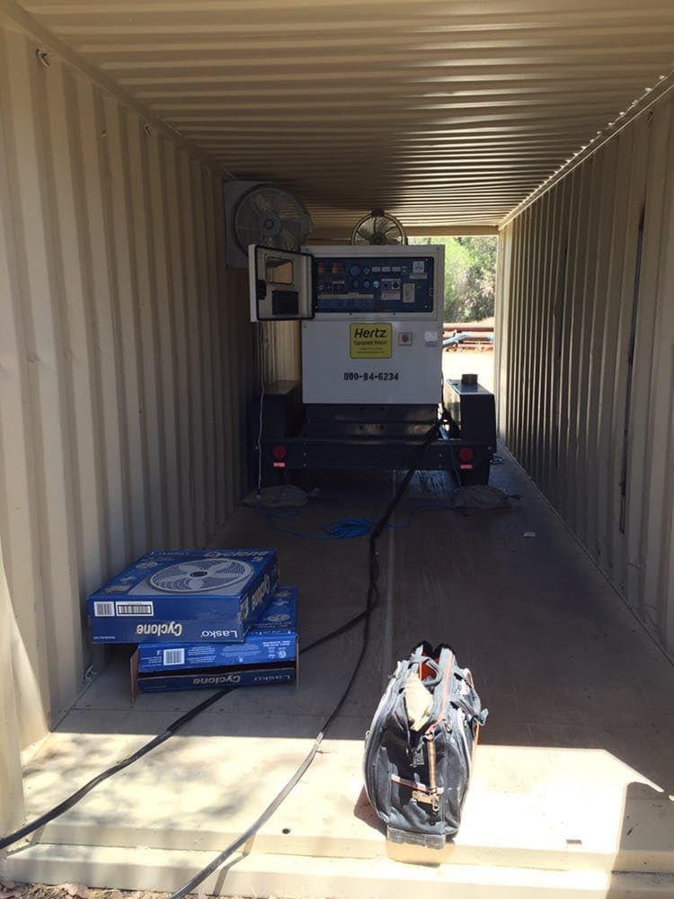 A mobile generator sits inside a metal shipping container with a tool bag and boxes on the floor in the foreground.