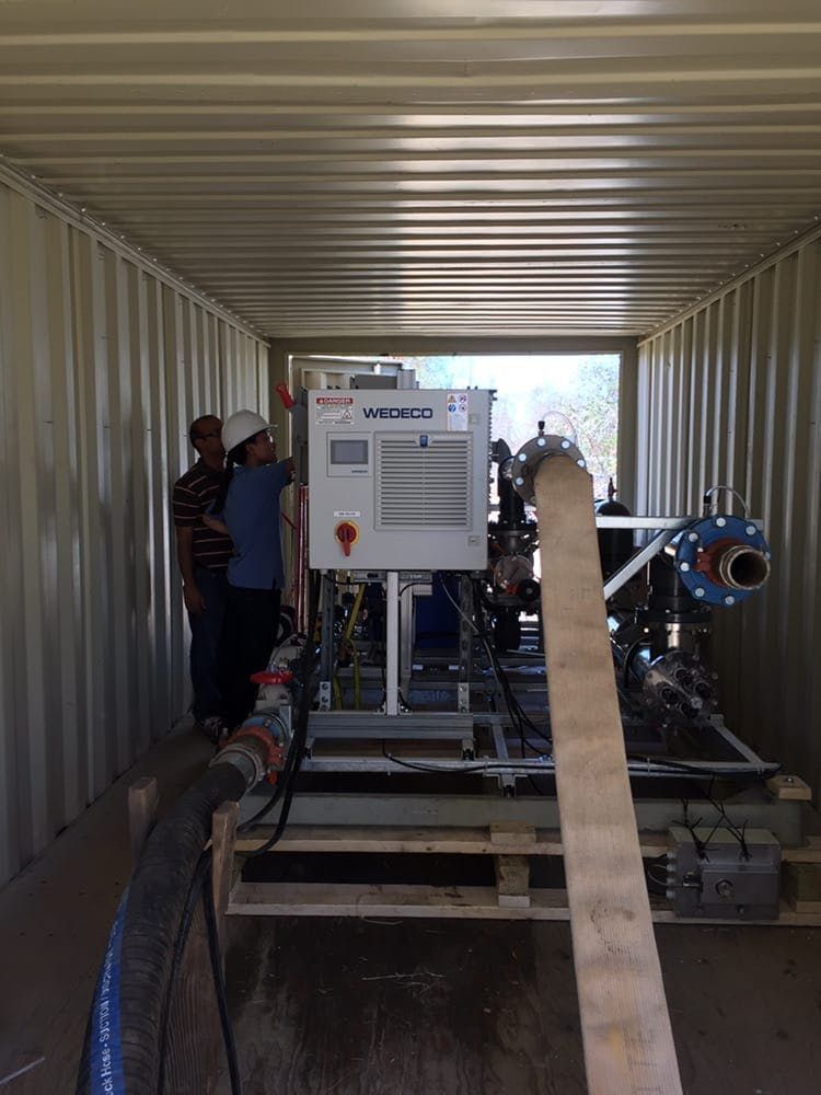 Two people examine a white industrial control unit and piping inside a shipping container.