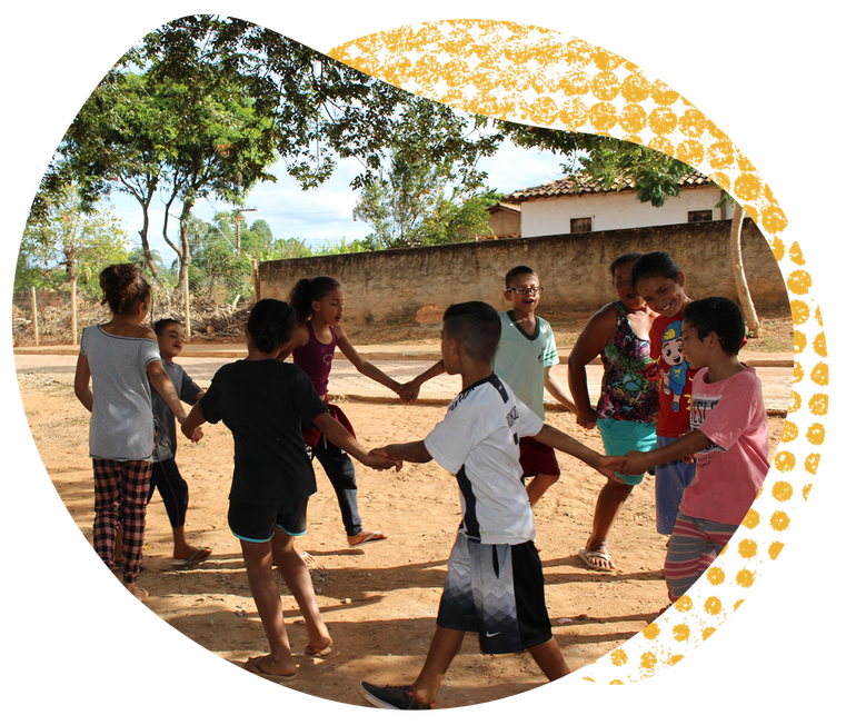 Um grupo de crianças vestidas casualmente dá as mãos em círculo num parque infantil de terra batida, brincando ao ar livre.