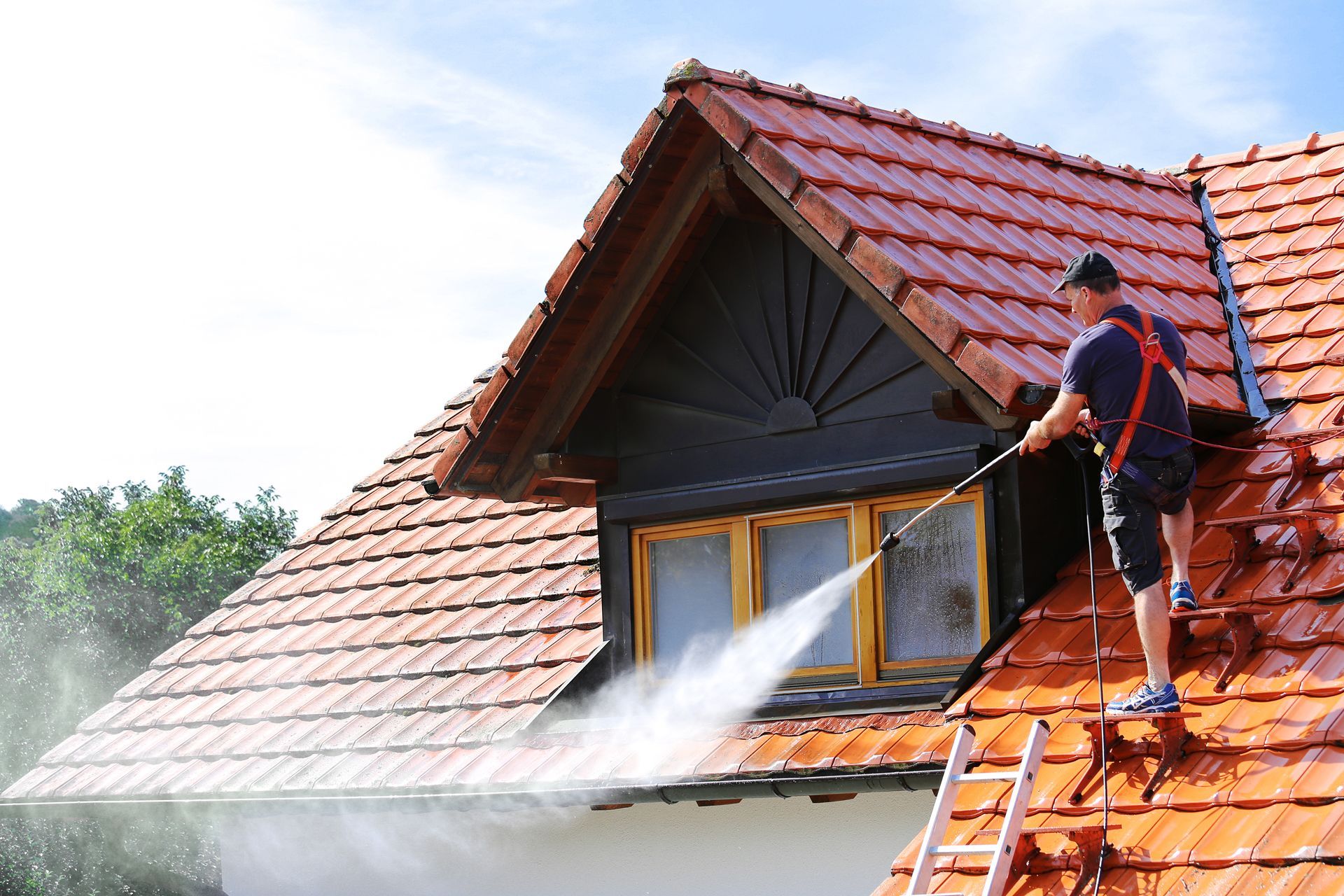Worker pressure washing a red-tiled roof on a house from a ladder