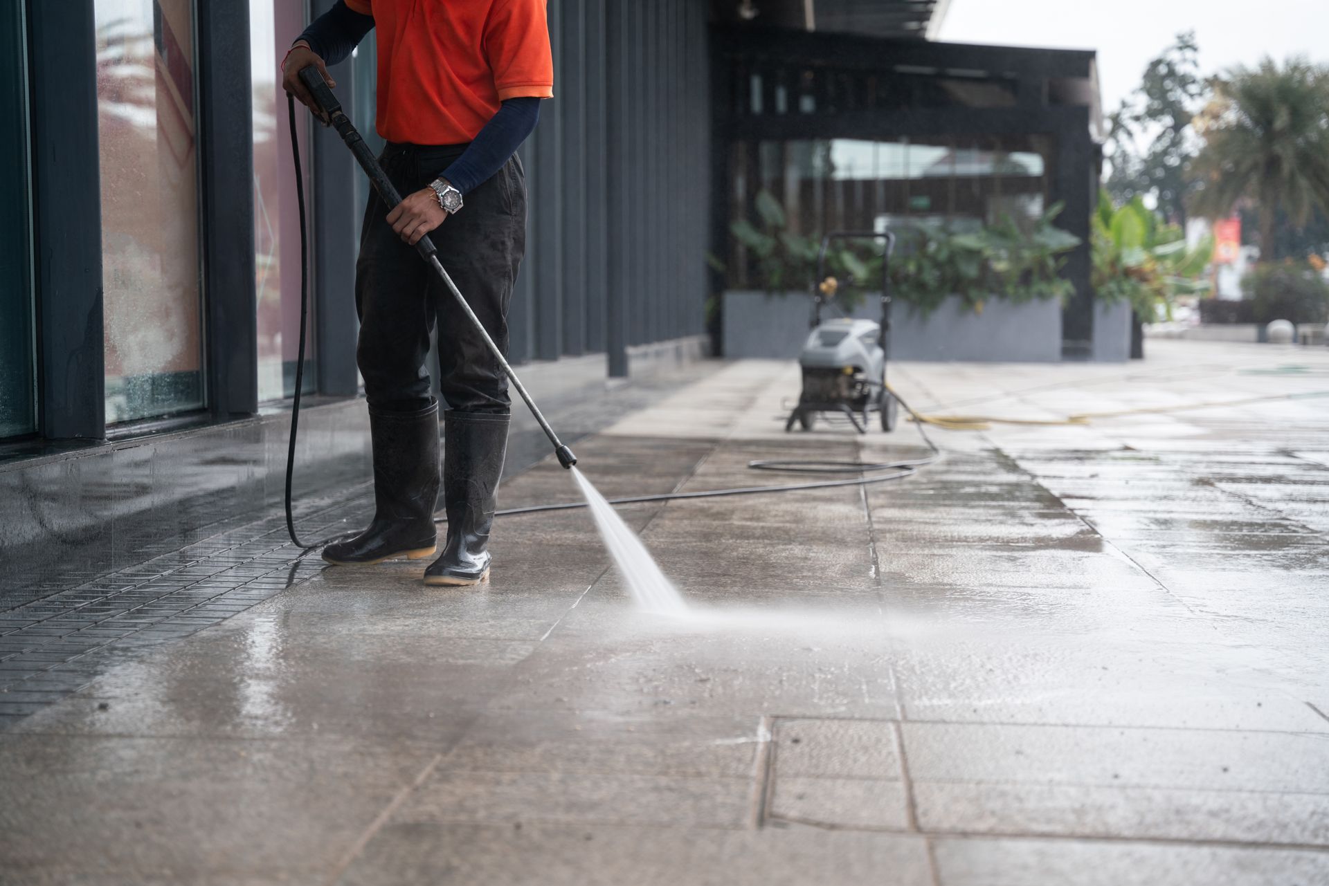 Worker pressure-washing a sidewalk outside a storefront, spraying water across wet pavement.