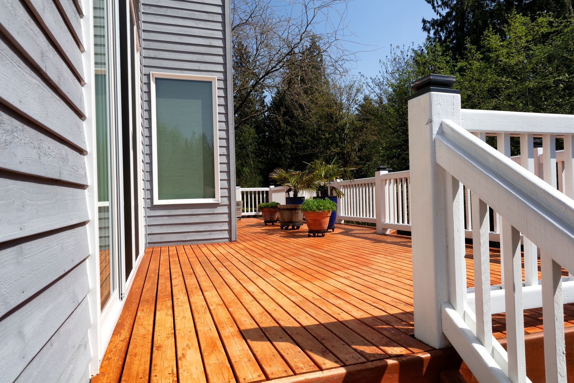 Wooden deck with white railing beside a house, featuring potted plants and trees beyond the patio.