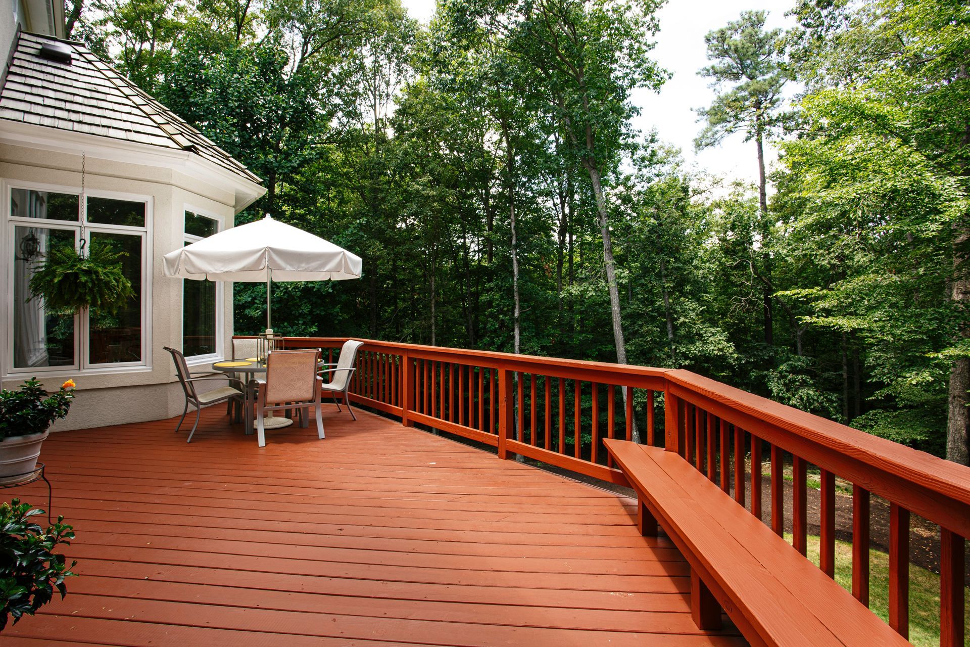 Wooden deck with railing beside a house, patio table, umbrella, and trees beyond.