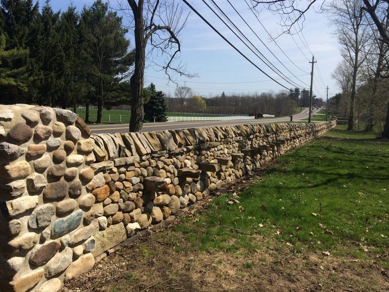 A stone wall surrounds a grassy field with a road in the background.