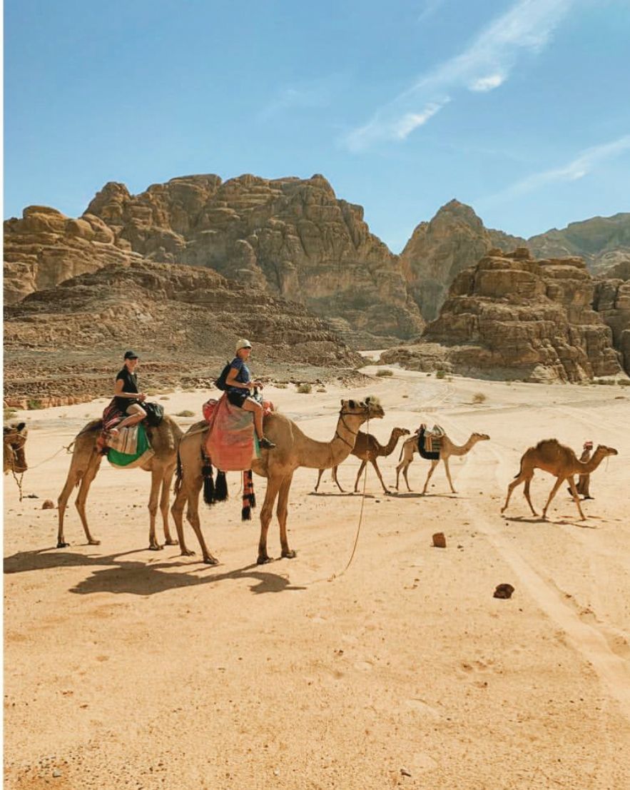 Twee mensen rijden op kamelen door een zanderige woestijn met rotsachtige bergen op de achtergrond. Andere kamelen lopen in de buurt.
