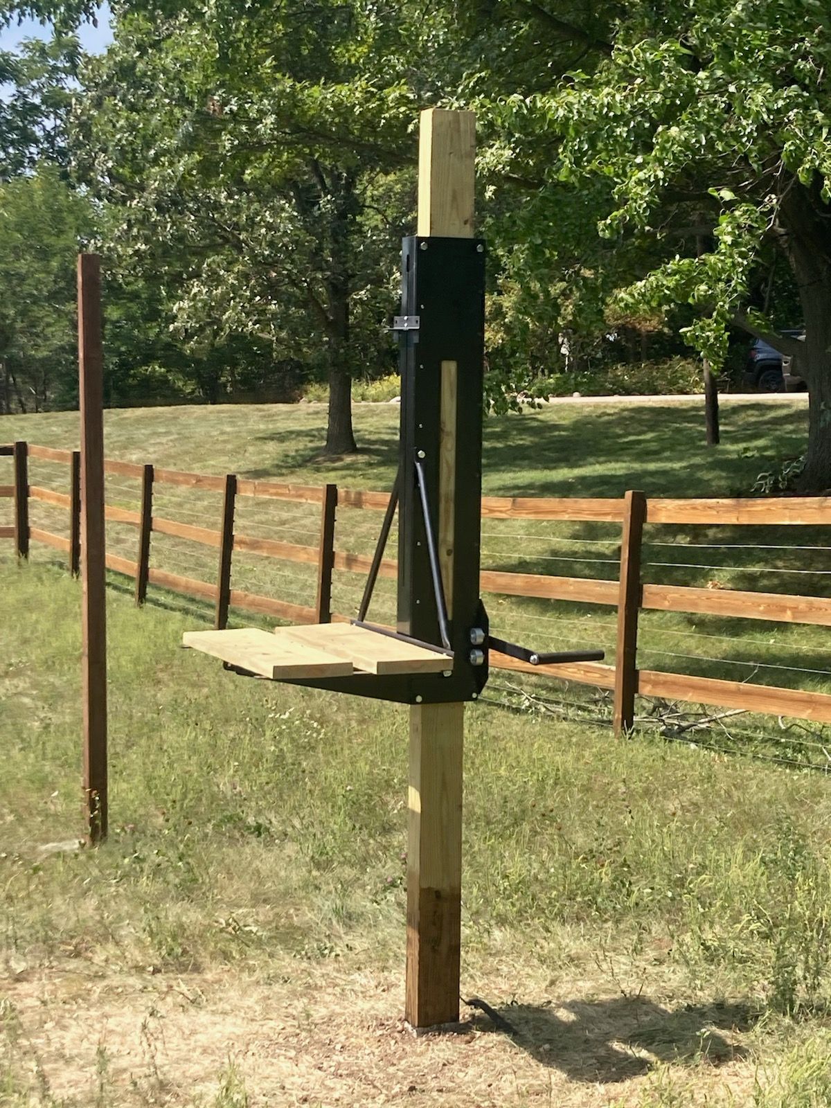 A wooden bench is sitting on top of a wooden post in a field next to a wooden fence.