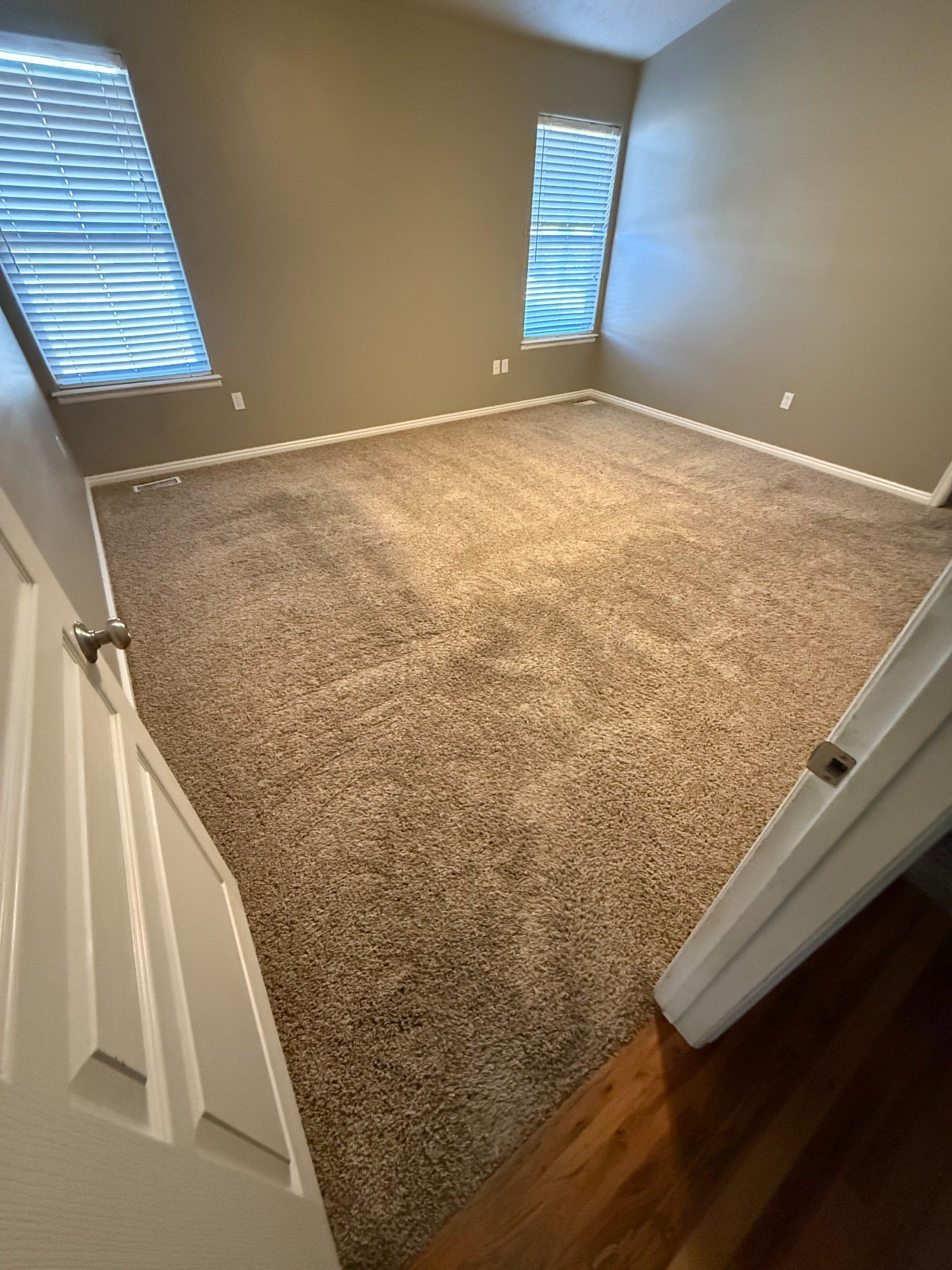 Empty beige carpeted bedroom viewed from doorway with two windows and white trim