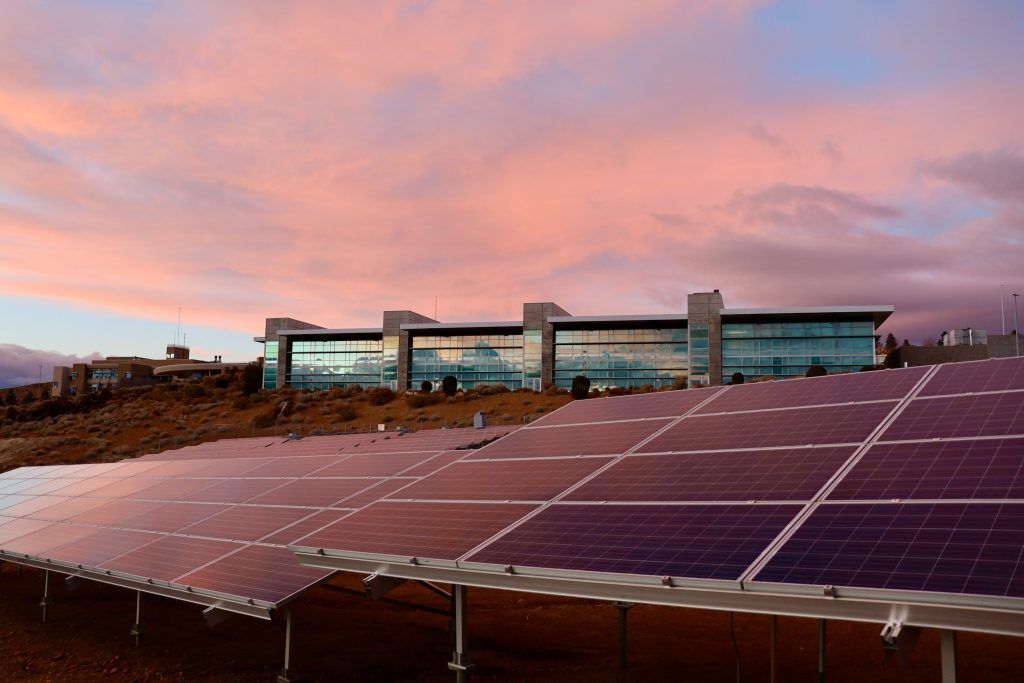 A row of solar panels are sitting in front of a building at sunset.