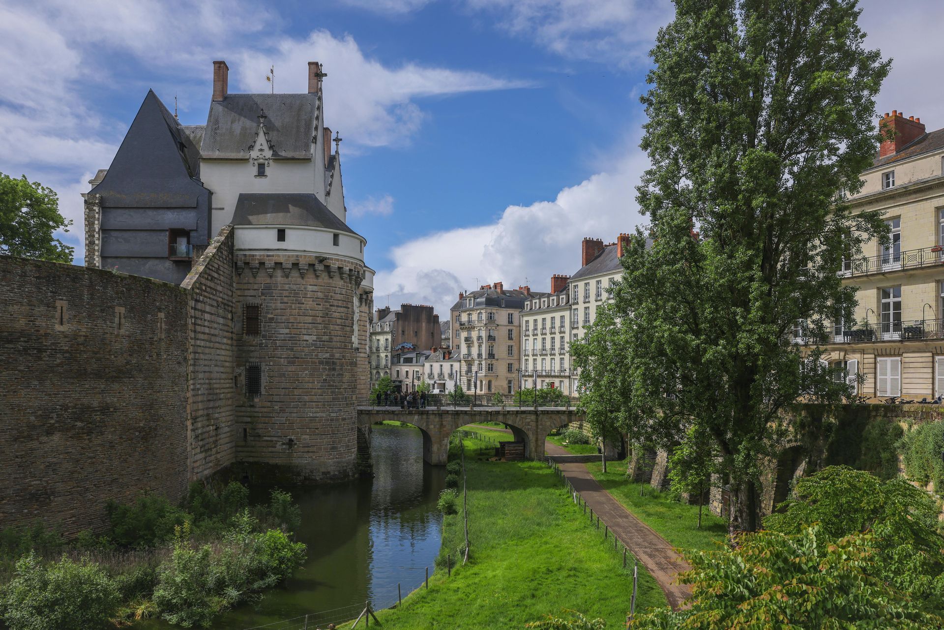 Château de pierre avec douves d'eau, herbe verte et pont dans une ville européenne sous un ciel bleu.