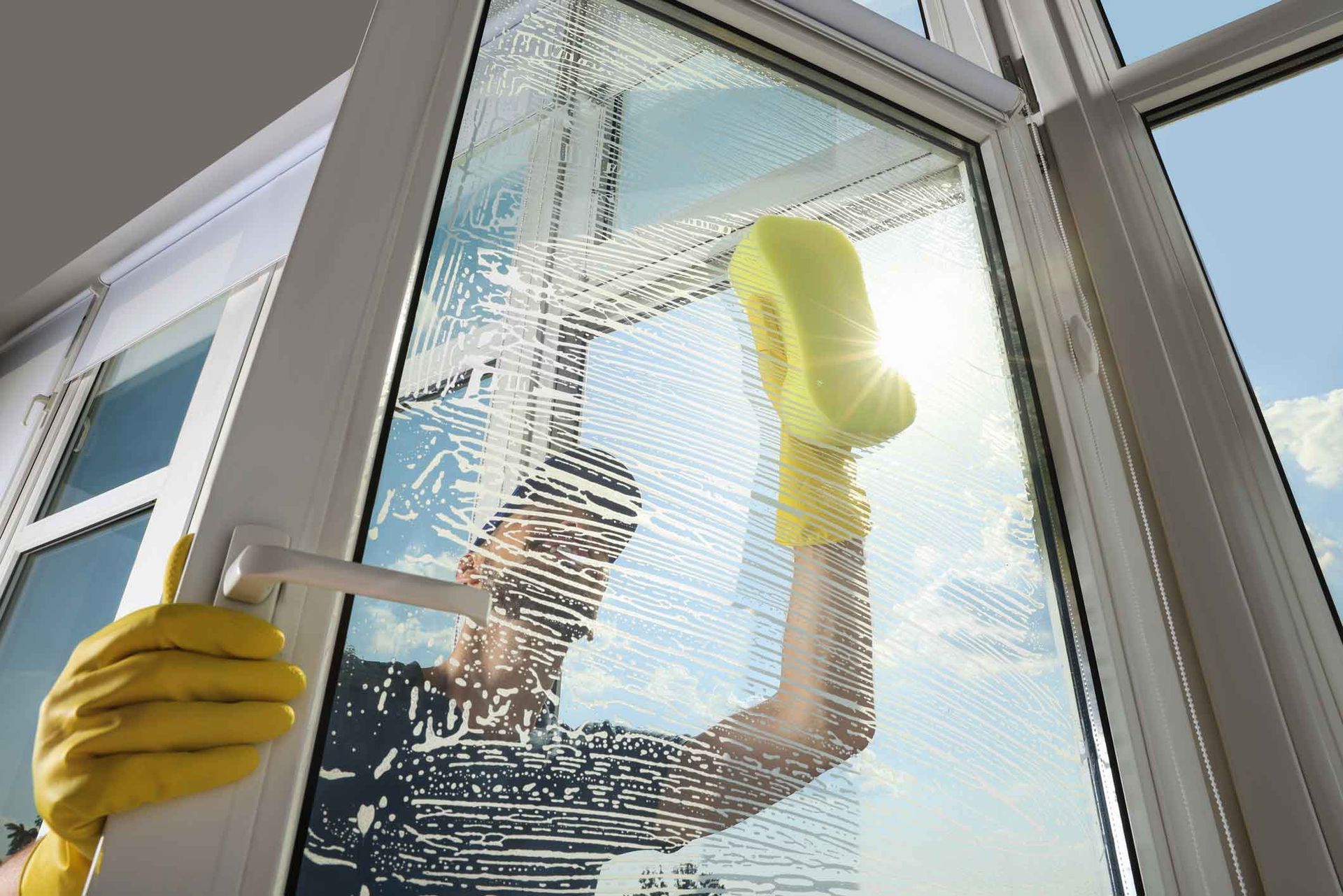Person wearing yellow gloves cleans window with a sponge, using soapy water, outside view visible.