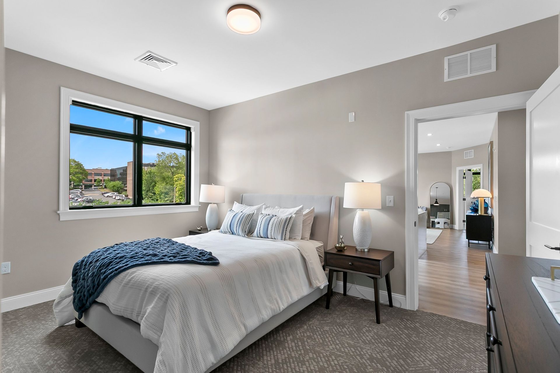 Bright bedroom with a neatly made bed, bedside lamp, and large window overlooking trees and rooftops.