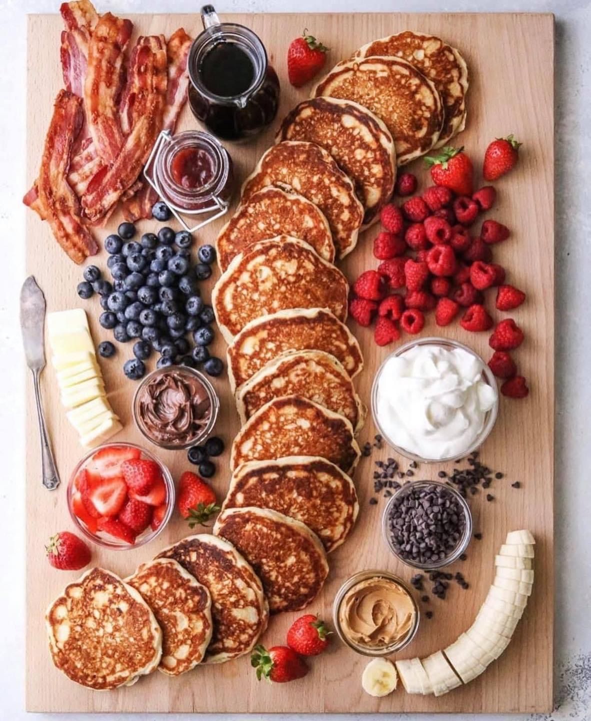 Breakfast charcuterie board with pancakes, bacon, fruit, and toppings.