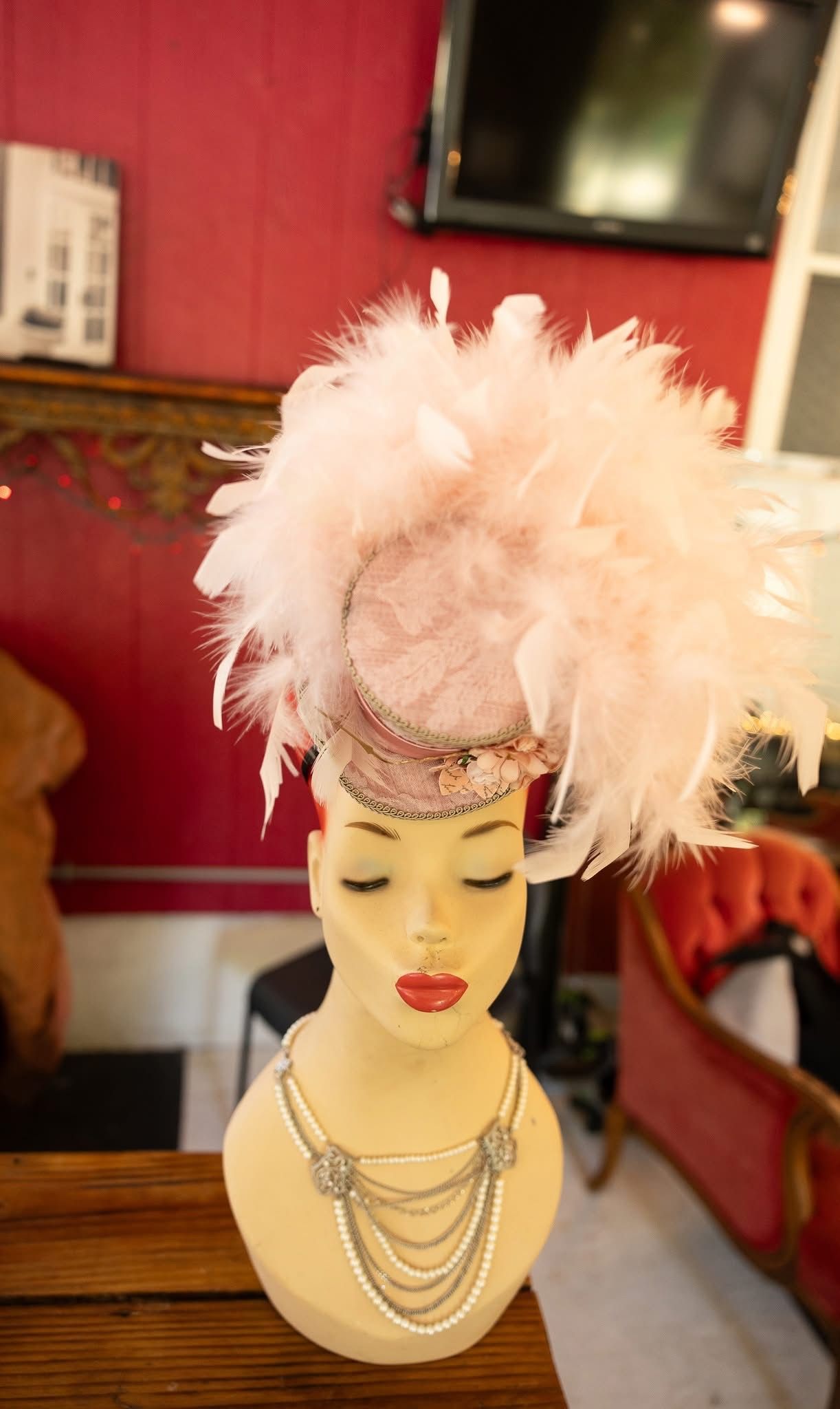 Pink feathered hat on a mannequin with a pearl necklace, set against a red wall.