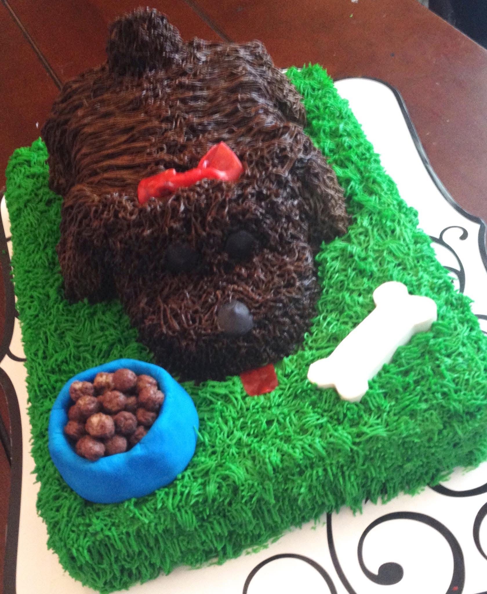 Dog-shaped chocolate cake with green grass frosting, red bow, food bowl, and bone.
