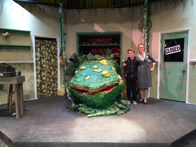 Two people pose with a large, green puppet plant on a stage set. Little Shop of Horrors