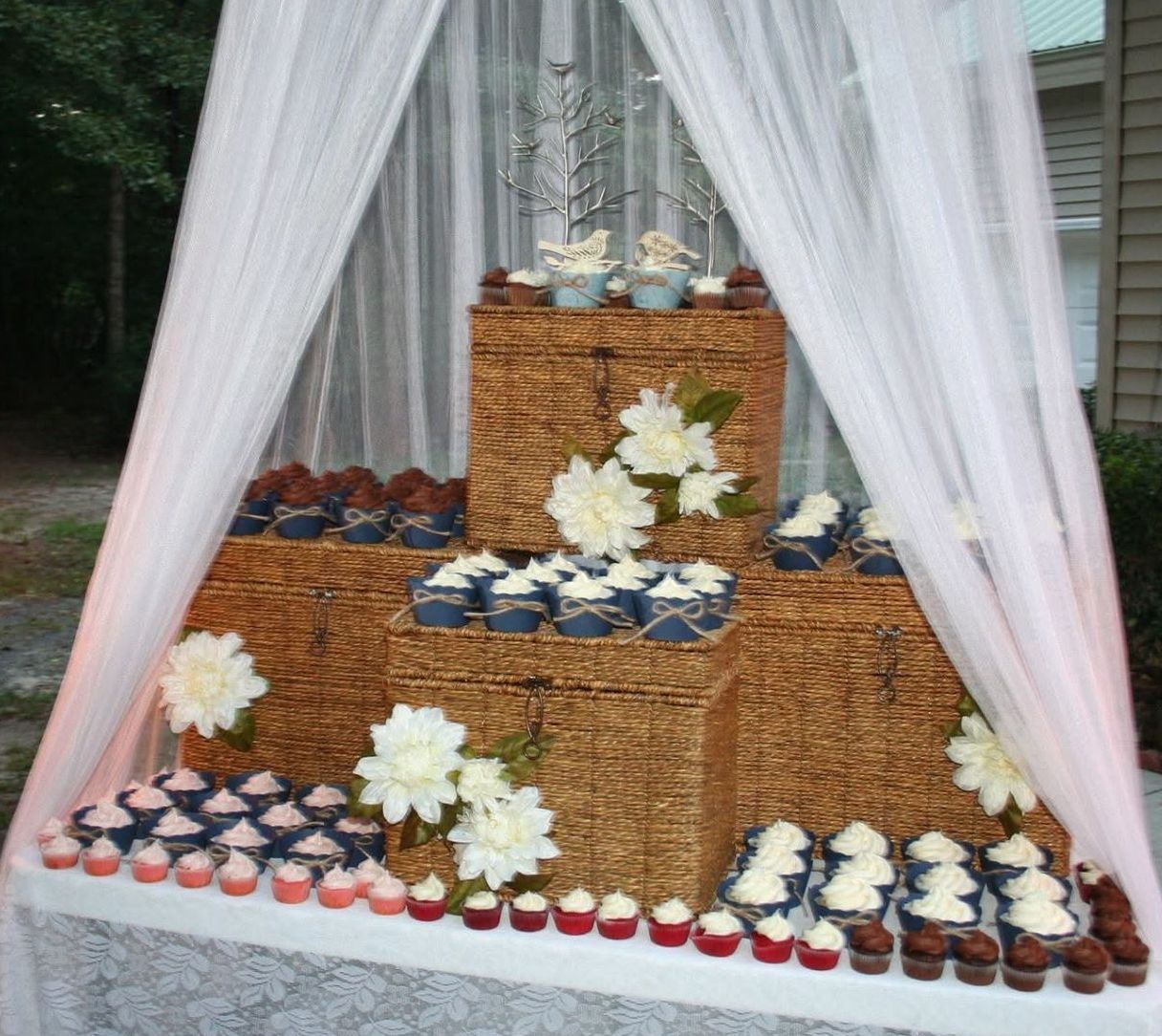 Cupcake display on stacked wicker boxes, decorated with flowers and white draping.