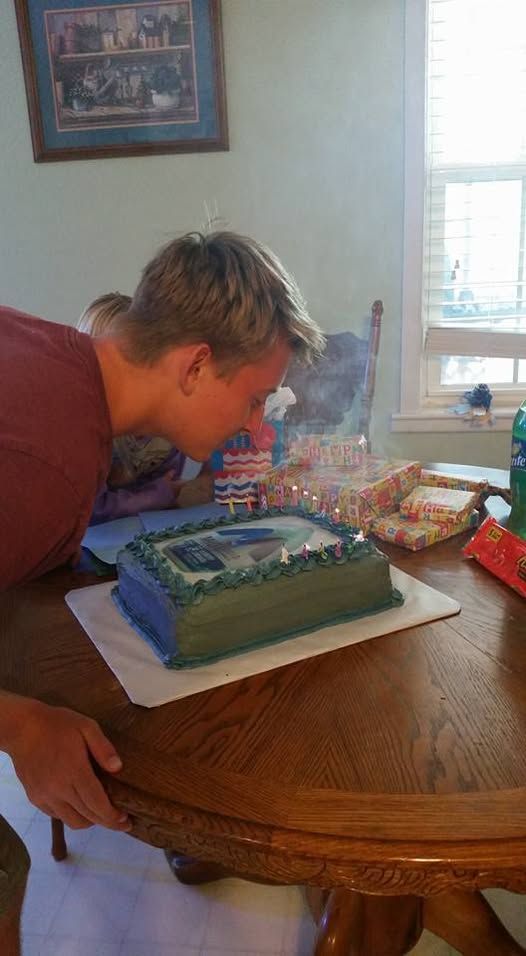 Man blowing out candles on a birthday cake at a wooden table in a brightly lit room.