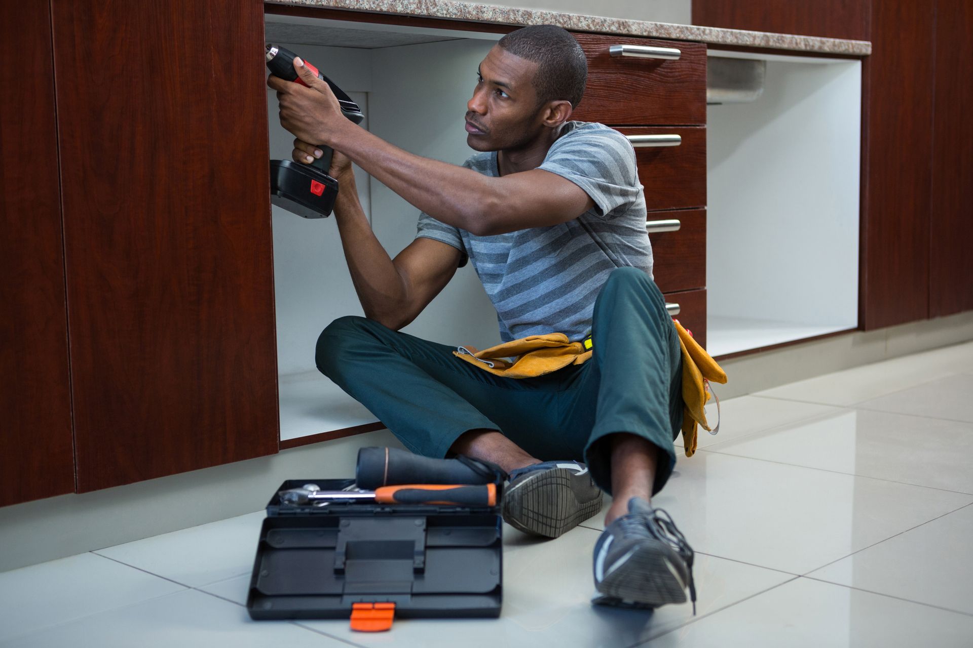 A man is sitting on the floor fixing a sink in a kitchen.