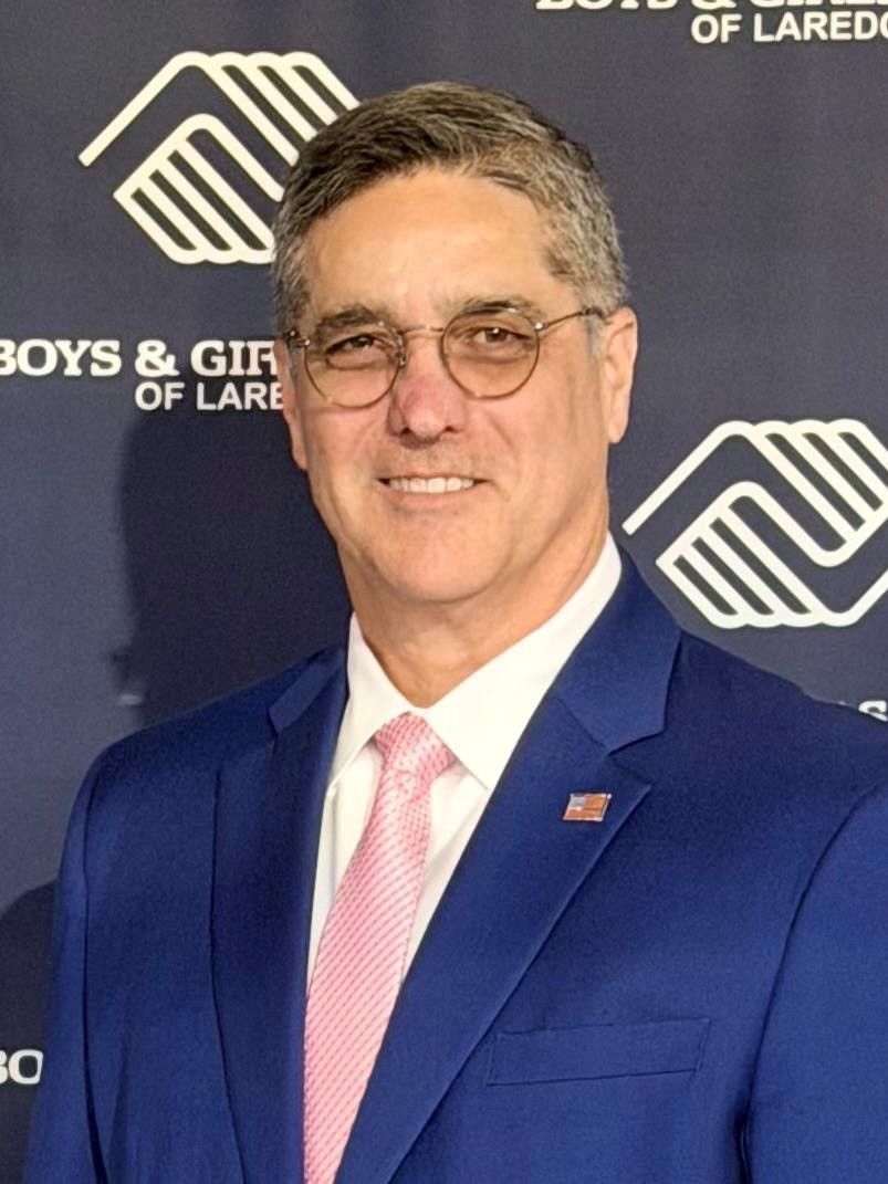 Football coach with arms crossed, standing on a football field. He wears a navy polo shirt and gray pants.