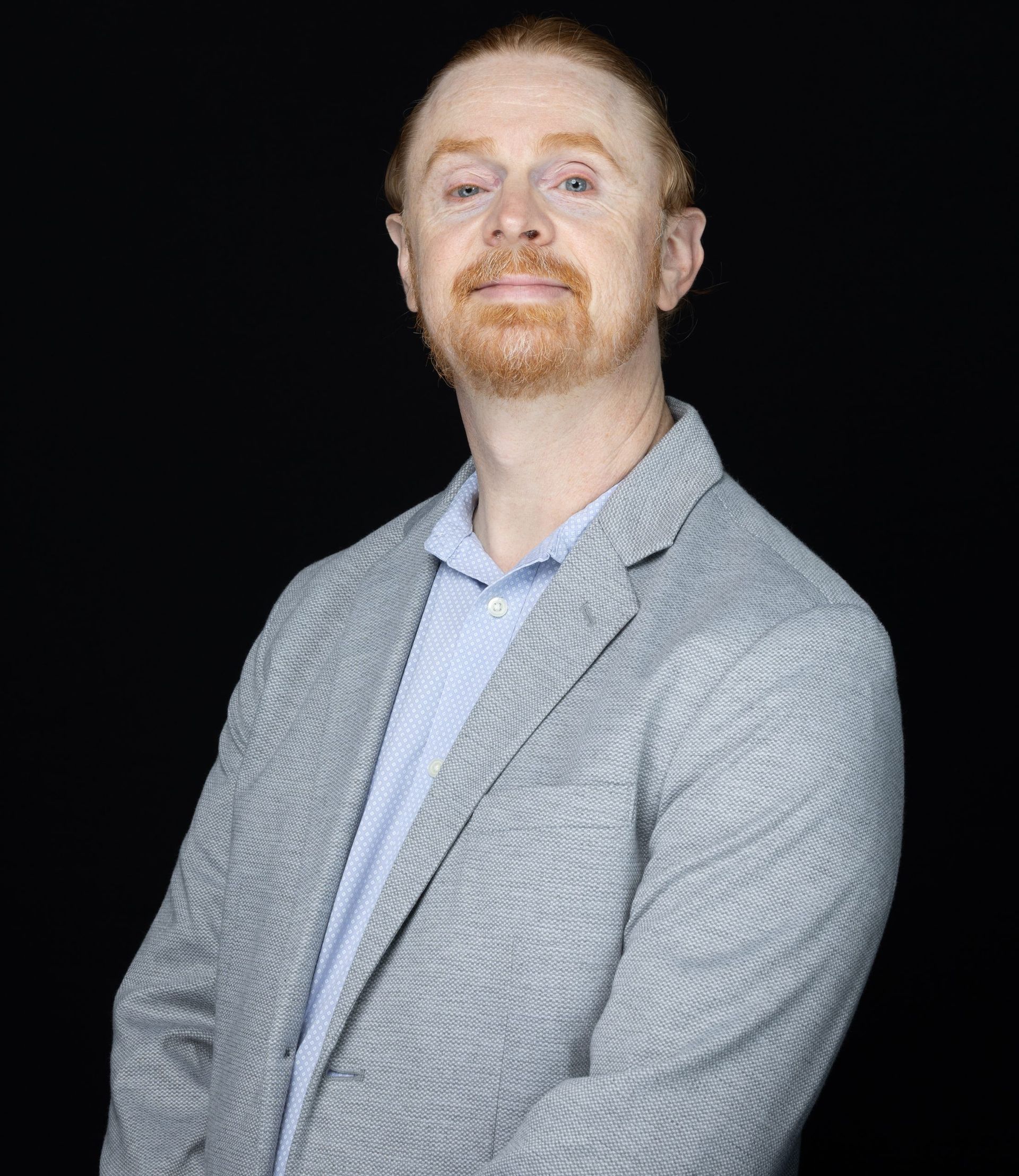 Football coach with arms crossed, looking stern on a sunny field, players in the background.