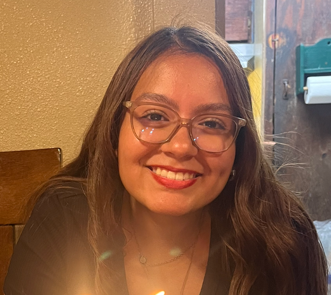 Football coach with arms crossed, looking stern on a sunny field, players in the background.