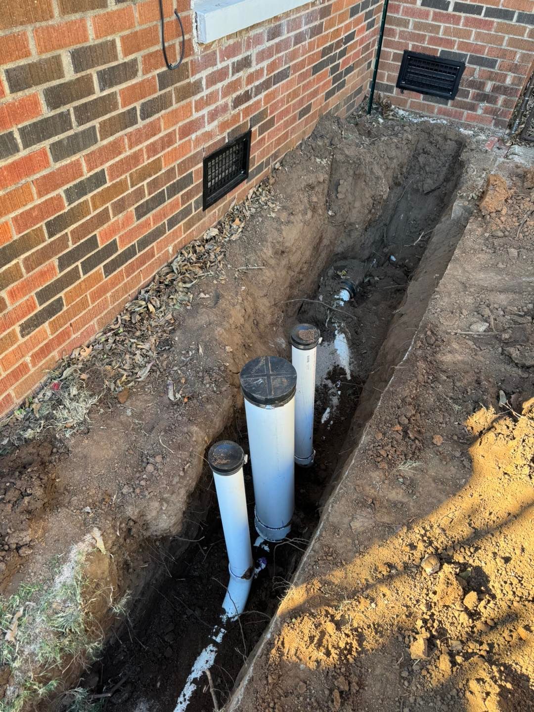 A drain pipe is being installed in the dirt in front of a brick building.
