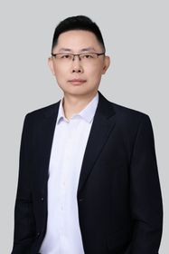 Man in grey blazer smiling, seated indoors near a window.