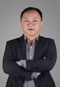 Man in grey blazer smiling, seated indoors near a window.