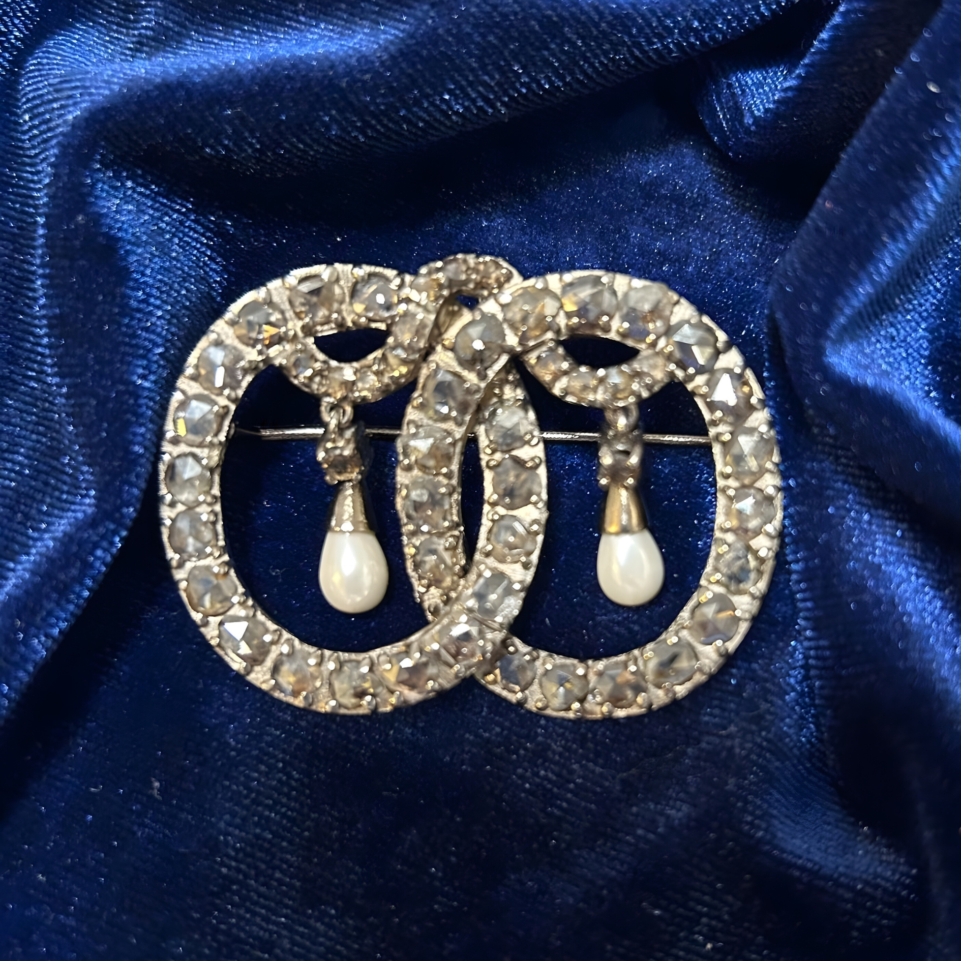 A silver-tone double-ring brooch set with rhinestones and two dangling pearl-like drops, displayed on blue velvet.