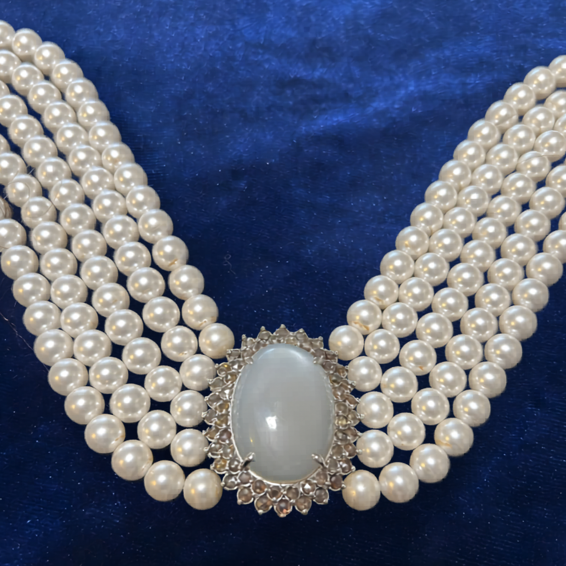 A multi-strand pearl necklace with a central, oval, milky-white cabochon gemstone framed by crystals, on blue velvet.