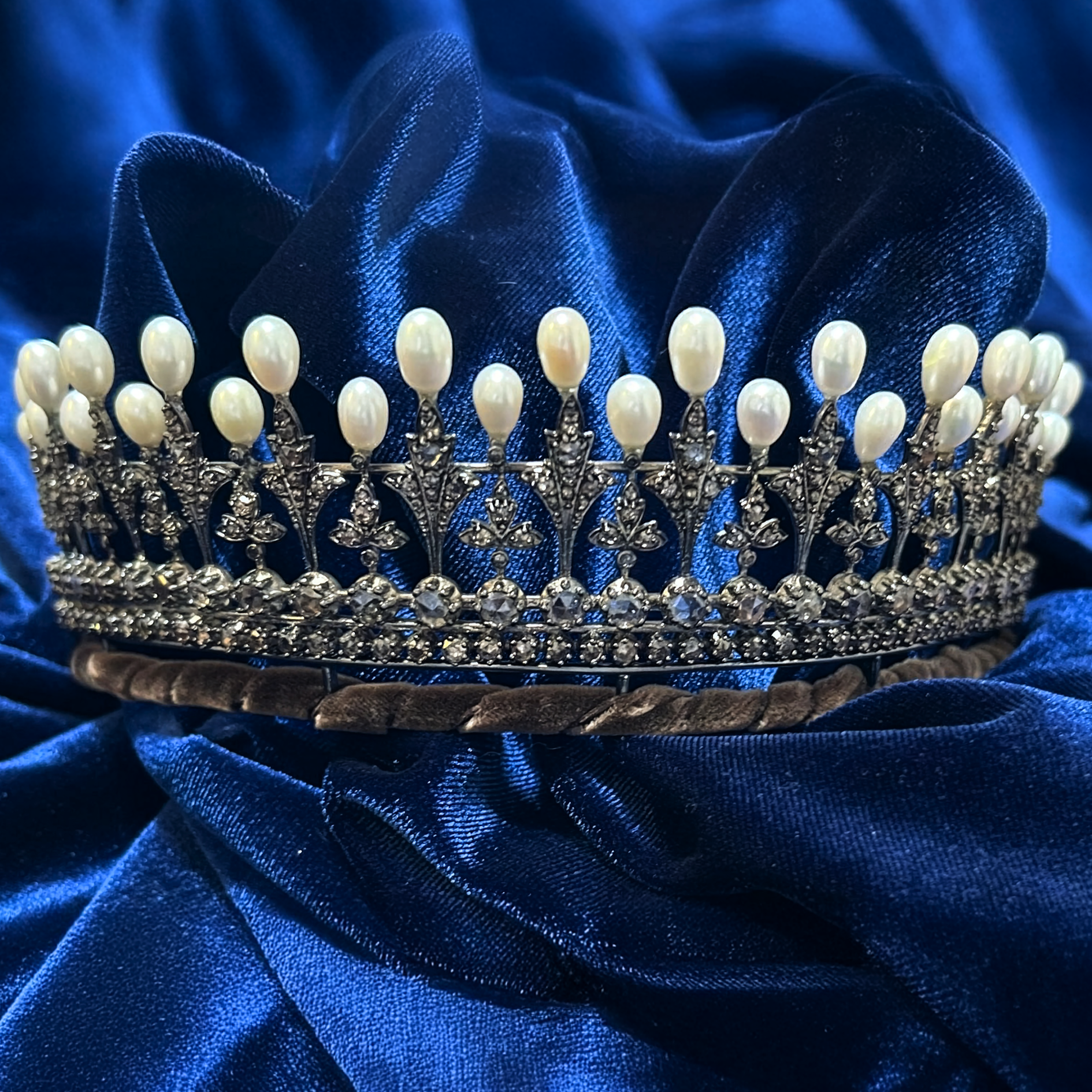 A silver, diamond-encrusted tiara topped with a row of drop-shaped pearls, displayed against a dark blue velvet background.