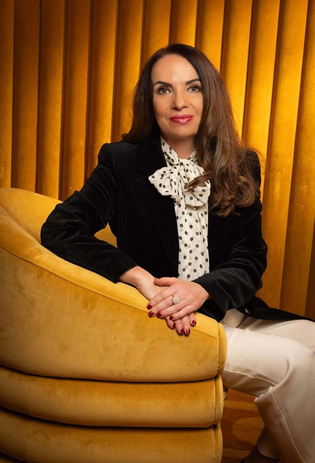 A person in a dark velvet blazer and polka-dot blouse sits in a curved yellow chair against a golden pleated background.