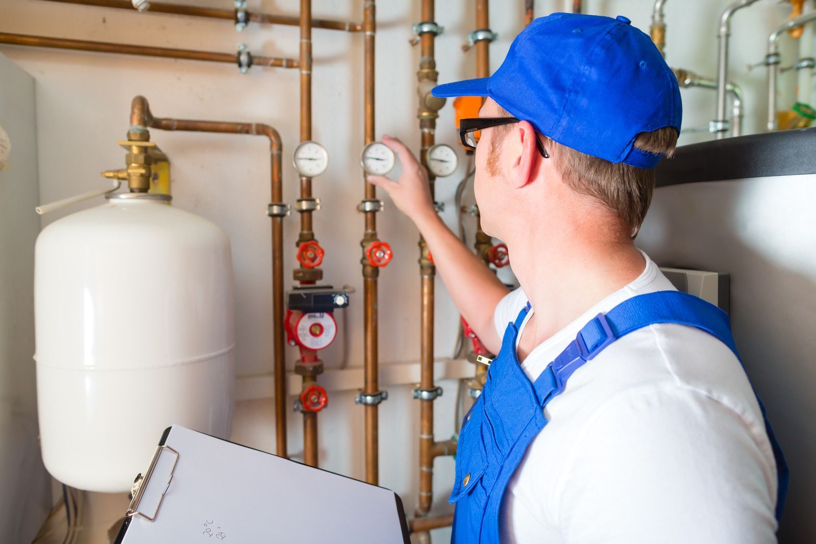 A man in a blue hat is working on a pipe while holding a clipboard.