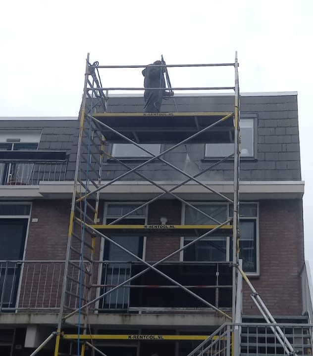 Persoon op een steiger die op een bewolkte dag het grijze dak van een bakstenen gebouw schoonmaakt.