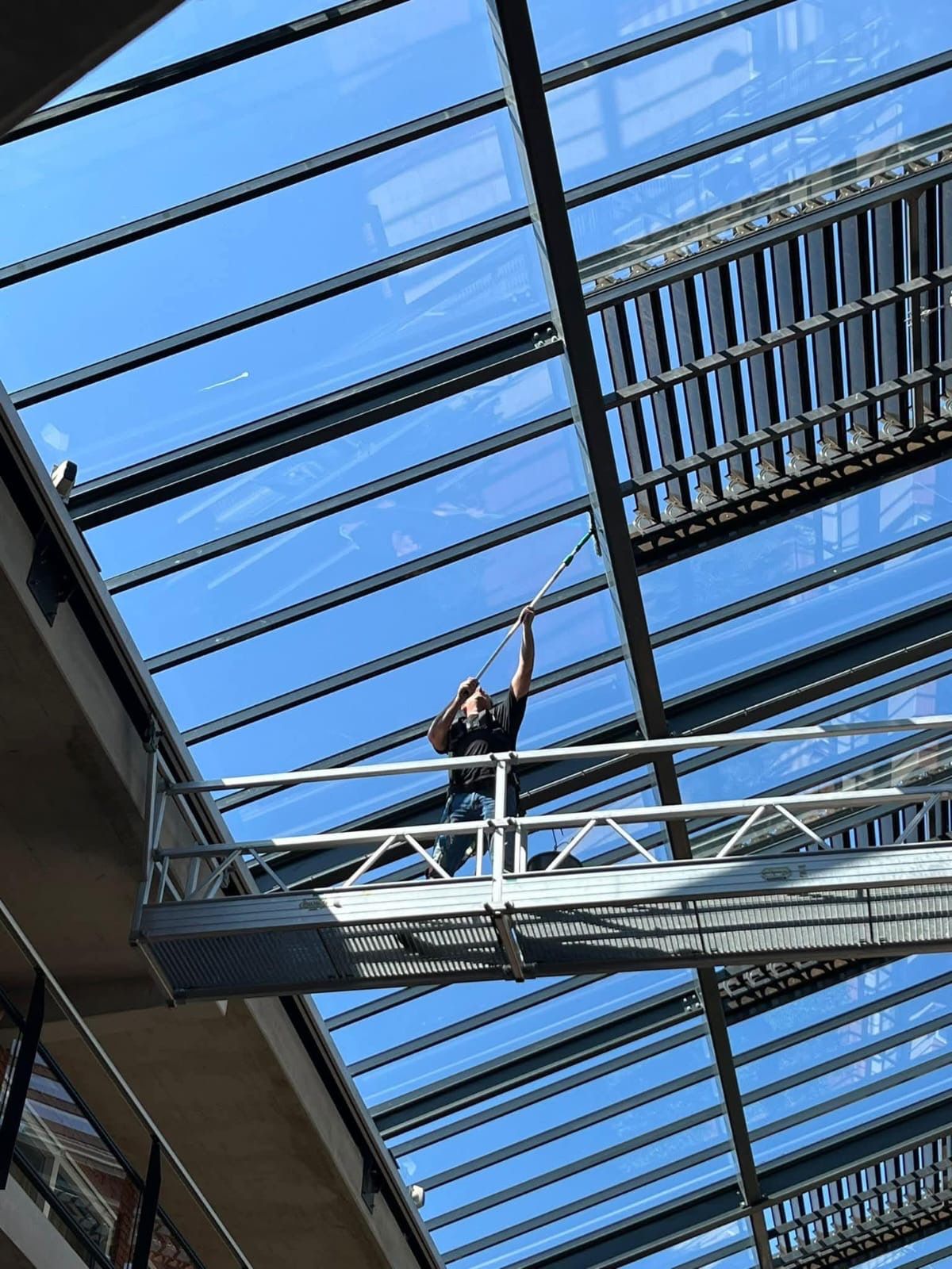 Een persoon op een platform maakt een glazen plafond schoon met een gereedschap met een lange steel, onder een blauwe lucht.