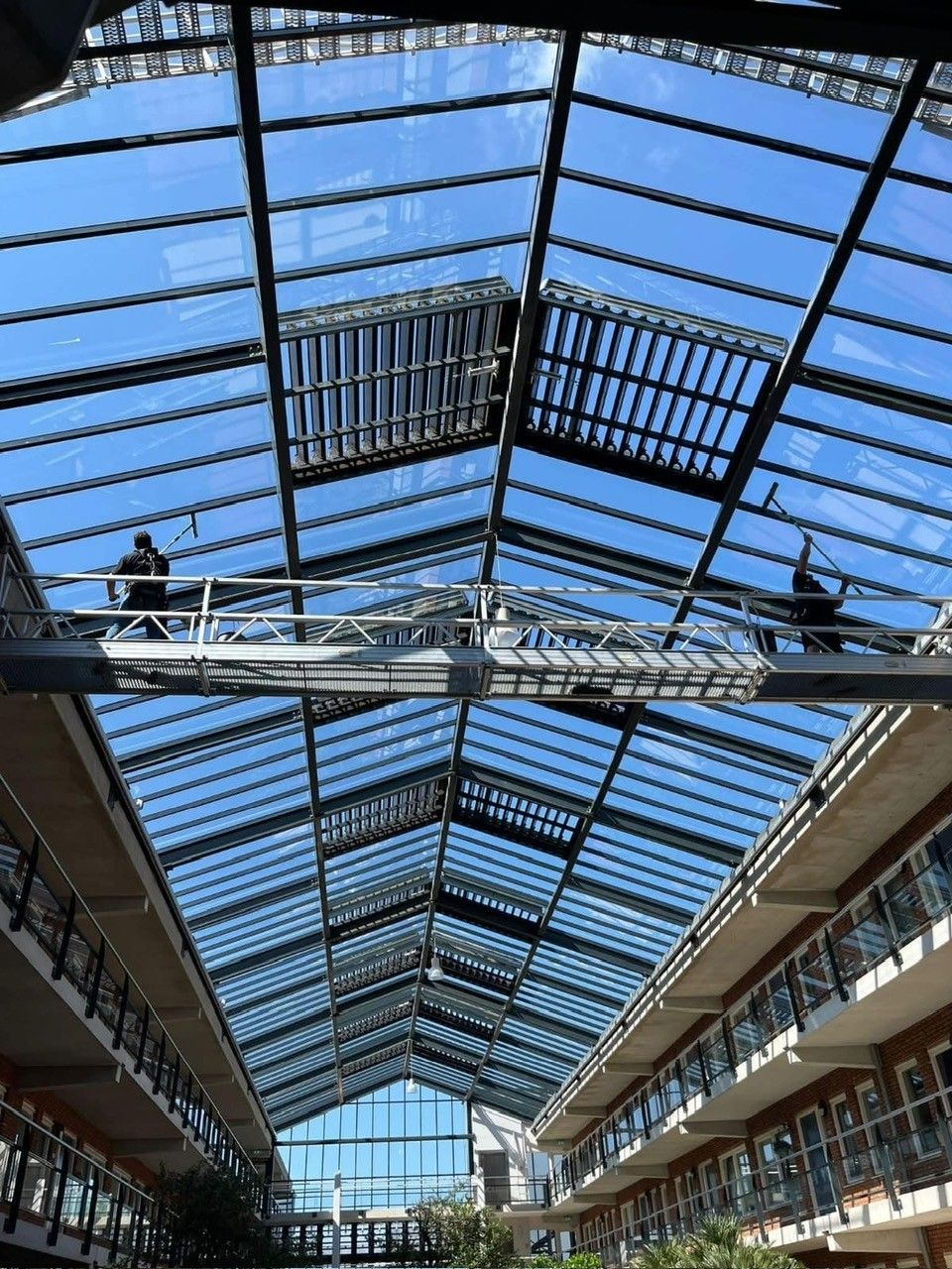 Werknemers op een glazen dakconstructie, met een heldere hemel erboven. Modern gebouw met loopbruggen.