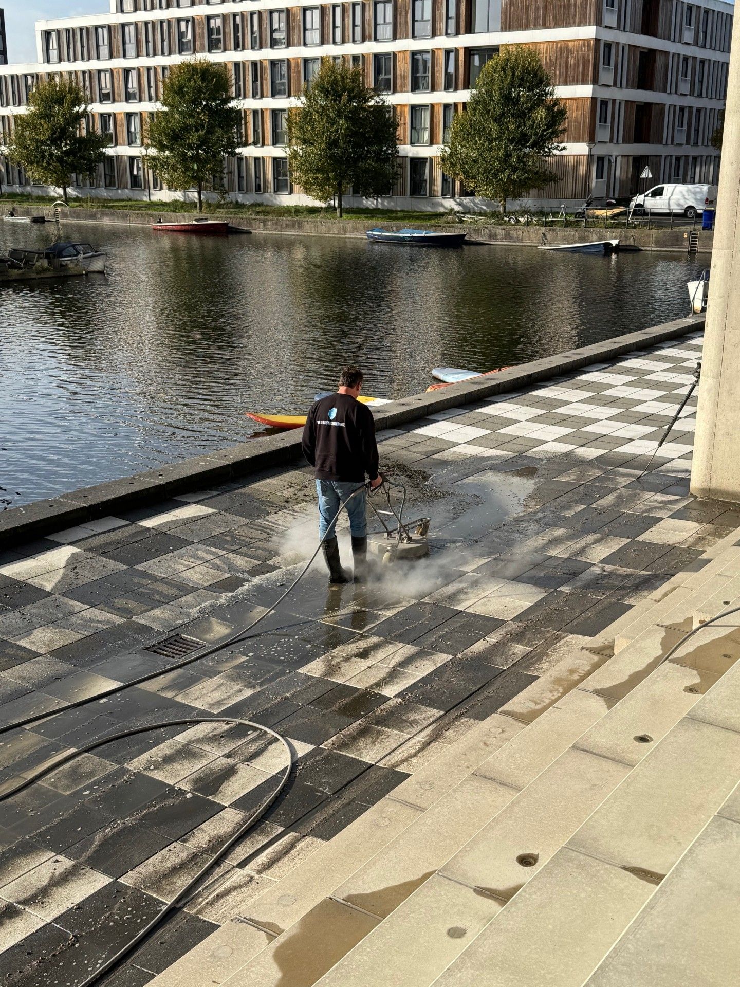 Met mankracht wordt een aangelegd wandelpad langs een waterpartij schoongemaakt, met gebouwen op de achtergrond.